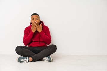 Young latin man sitting on the floor isolated shocked covering mouth with hands.