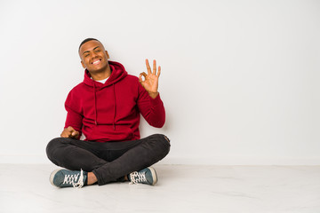 Young latin man sitting on the floor isolated winks an eye and holds an okay gesture with hand.