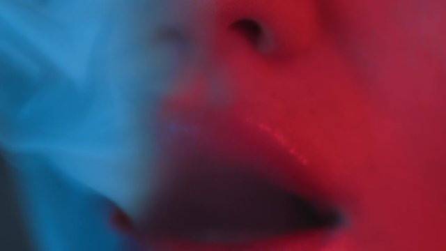 macro close up of mouth and shiny female lips exhaling cigarette smoke in neon lights, a woman or girl slowly smoking tobacco or drugs at party or night club