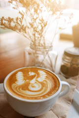 Hot latte art in white cup and dry flowers decoration in glass in natural sunlight at cafe