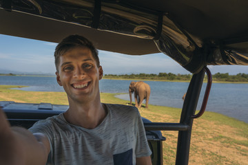 young man taking a selfie from a safari jeep with an asian elephant in the background