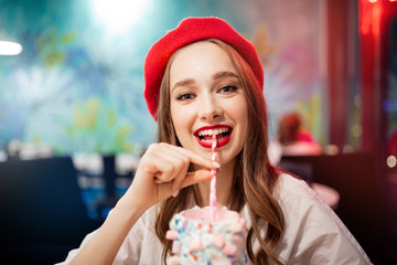 Girl with white teeth, beautiful smile in red beret, France drinks pink strawberry milkshake from...