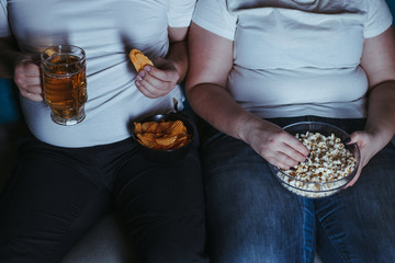 Relationship problems, emotional eating, laziness. Overweight couple watching tv eating junk food