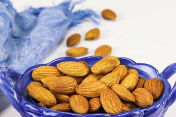 Almond nuts lie in an oval-shaped blue glass bowl and on a white surface, close-up