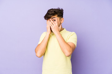 Young arabian man isolated on a purple background blink through fingers frightened and nervous.