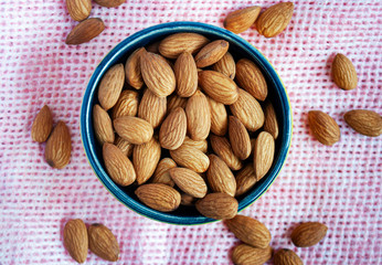 Dried almonds in a bowl surrounded by another almonds on a pink surface.