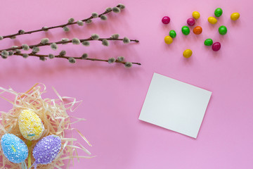 Colorful easter eggs in a hay nest, willow branch, sweets and a blank card on the pink background. Easter holidays concept. Copyspace, place for text and wording. April vibes.
