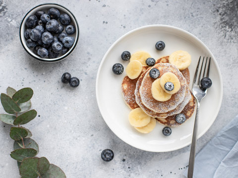 pancakes with fresh blueberries, banana in a white plate on a gray background with a branch of eucalyptus. healthy food concept. horizontal image, top view, flat lay
