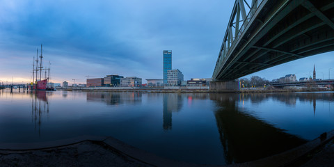 long exposure panorama of the Überseestadt in Bremen, Germany with office building, sail boat and perfect reflection on the river weser during blue hour, last light hitting the commercial district