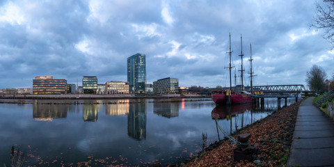 Naklejka premium panorama of the Überseestadt in Bremen, Germany with office building, sail boat and perfect reflection on the river weser during blue hour