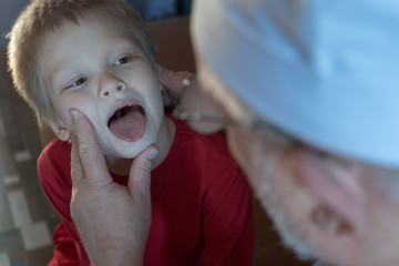 Home doctor in white coat, cap and glasses with gray beard examines two children teenagers at home....