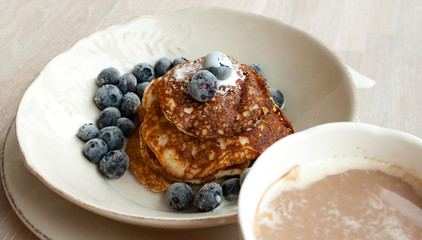 Low Carb Keto Diet Pancakes from almond coconut flour with blueberries, cream on white plate and cocoa cup on wooden table background close up view. Selective focus. Copy space. Ketogenic concept