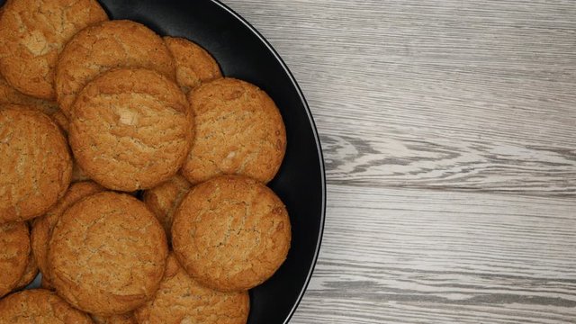 Top View Rotating Close Up Of Fresh Brown Oatmeal Cookies On A Black Plate On A Gray Wooden Background On The Left Side.