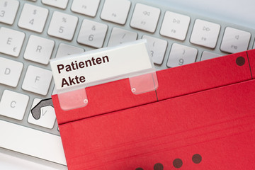 Roter Hängehefter mit Reiter und dem Text: Patienten Akte. Der Ordner liegt auf einem Computer...