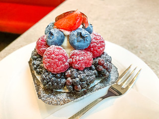 Cream cheese berry tartlet on a white plate with fork. Cream cheese in tart shell, garnished with blackberries, raspberries, blueberries and strawberries on top, dusted with icing sugar. Food photo.