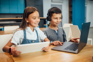 Two happy small friends, boy and girl using smart digital tablet at modern blue classroom.