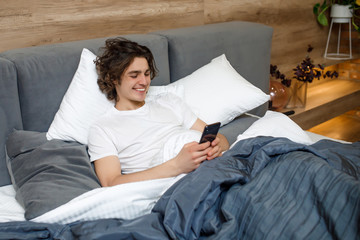 Handsome young man in pajamas using a cellphone while lying in bed in the morning