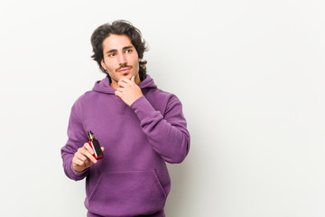 Young man holding a vaporizer looking sideways with doubtful and skeptical expression.