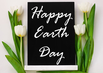 Happy Earth Day April 22 greeting message sign on black chalk board with tulip flowers on white background. World ecology concept card.Saving environment, save clean planet.Blackboard with text,banner