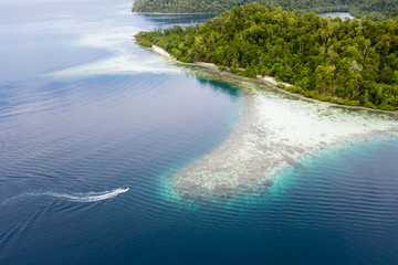 A small boat cruises along the edge of a coral reef in Raja Ampat, Indonesia. This tropical region...