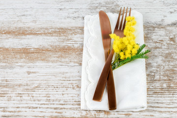 Table set of khife and fork with a branch of spring yellow mimosa on the white napkin and on the natural wooden table. Vintage, holiday concept.