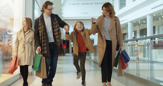 Caucasian family with two kids spending weekend in shopping mall. Smiled parents walking hand in hand with small son daughter with bags from shop. Little children having fun with mother and father.