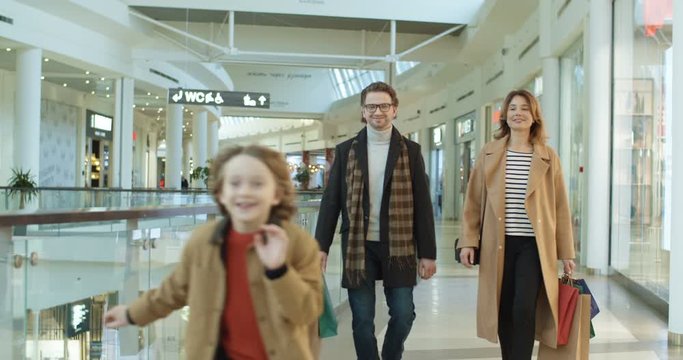 Caucasian family with little son spending weekend in shopping mall. Happy parents walking hand in hand with small boy carrying bags from shop. Kid running away mother and father.