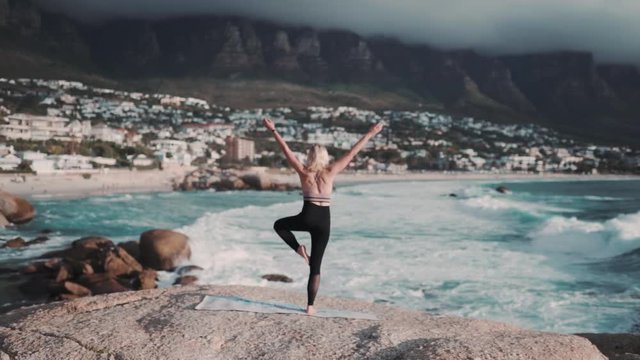 Beautiful and healthy female doing a yoga pose in front of a amazing blue seascape with misty mountains in the back.