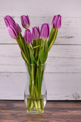 Purple tulips in clear vase on wood base with light wood background in the center of page 