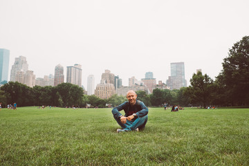 Naklejka na ściany i meble Happy man sitting on grass, enjoying vacation outdoors in Central Park, New York City, USA. Smiling young tourist man in summer sun relaxing in Manhattan, visiting NY.