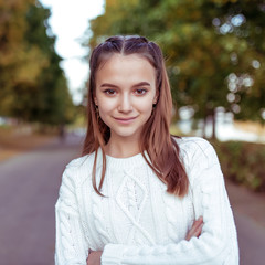 Close-up portrait of girl, teenager schoolgirl, in white knitted sweater, in summer in park. Happy smiles, emotions of positive and joy. Long hair. Rest after school, background trees.