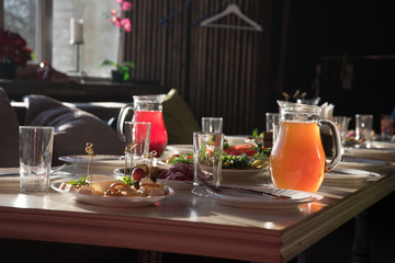 Table setting for a party or Banquet in a cozy small room. There are dishes and drinks on the table, and sofas with pillows are waiting for guests.