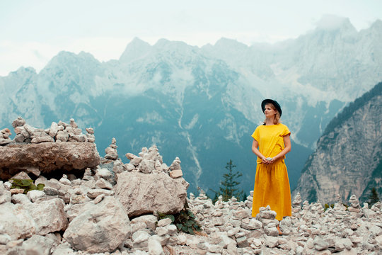 Portrait of a young woman in yellow standing in front of a  big mountain ridge with stone sculptures and holding a stone