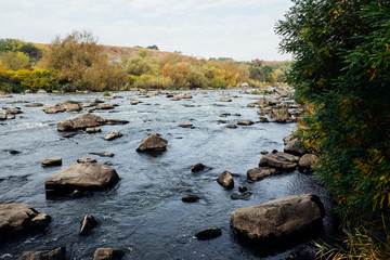 Clear blue river, lush green rural forest and rocky stone cascade in warm sunlight. Colorful autumn landscape, Southern Bug, Ukraine. Beautiful fall nature with green trees. Mountain hills view.