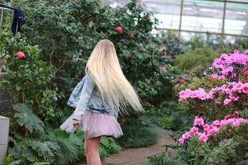 Young girl with long white hair in a denim jacket walks in the garden with blooming azalea