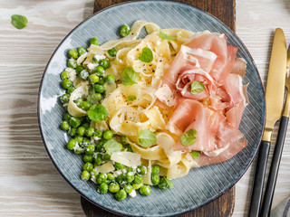 Linguine pasta with white sauce, green peas, Parmesan, prosciutto and fresh mint in a blue plate on wood board.