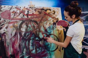 A young female artist is creating an abstract painting. She is working in the loft space with dimmed lights. She is using oil paint and a brush.