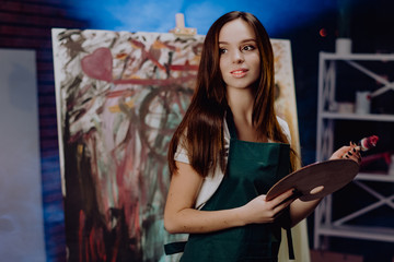 Obraz na płótnie Canvas A young female artist is creating an abstract painting. She is working in the loft space with dimmed lights. She is using oil paint and a brush.