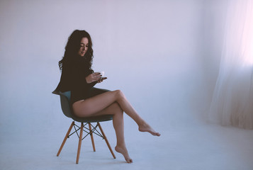 Fototapeta premium Beautiful woman drinking coffee sitting in chair wearing black elegant bodysuit in white room closeup. Good morning.