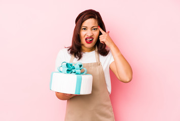 Middle age latin woman holding a cake isolated on a pink background showing a disappointment gesture with forefinger.