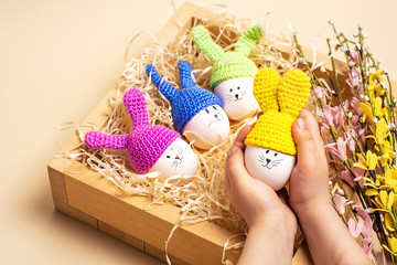 Easter background.Easter decorative eggs with painted cheerful faces and crocheted ears in the form of bunnies in hands of a child, spring flowers in wooden box on the beige background.DIY