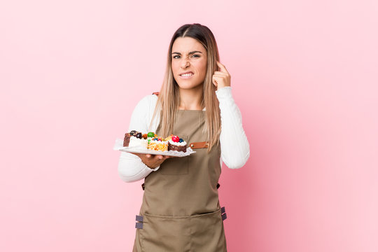 Young Baker Woman Holding Sweets Covering Ears With Hands.