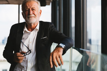 Retirement concept. Senior grey-haired businessman standing and looking to right hand at modern business lounge high up in an office tower.