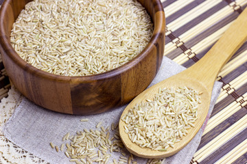 Uncooked brown rice in a bowl and spoon with light napkin on wooden background