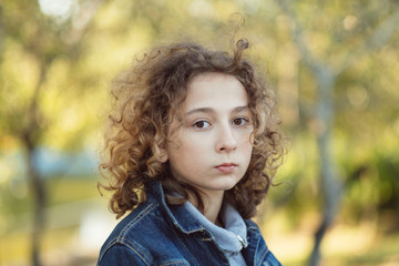 Offended and sad cute young girl outdoors with curly hair in the wind. Upset beautiful teenage child portrait on a windy day walking in the green park on the nature at summer or spring. 