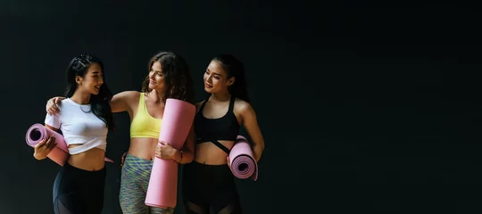 Fotobehang Panoramic banner. group of sporty multicultural women slim body smiling holding yoga mat at dark black wall background in fitness gym resting after yoga class, sport, yoga training, lifestyle concept © Vittaya_25