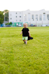 Happy boy running on green meadow. Little kid in black hero cloak running and smiling in the field on summer day outside in the park. Happy childhood concept. Cute child plays superhero outdoor.