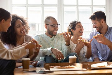 Five laughing multi-ethnic friends eating pizza indoors