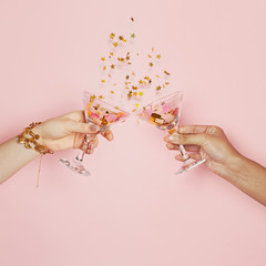 Celebration and holiday background. Two hands holding wine glasses with gold confetti on pink background