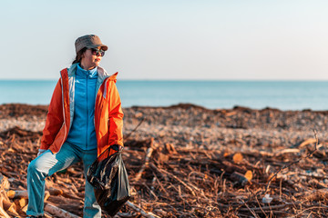 A young Caucasian female volunteer in a jacket, posing with a plastic garbage bag in her hands, posing against a dirty beach, sea and sky. Concept of environmental pollution. Copy space